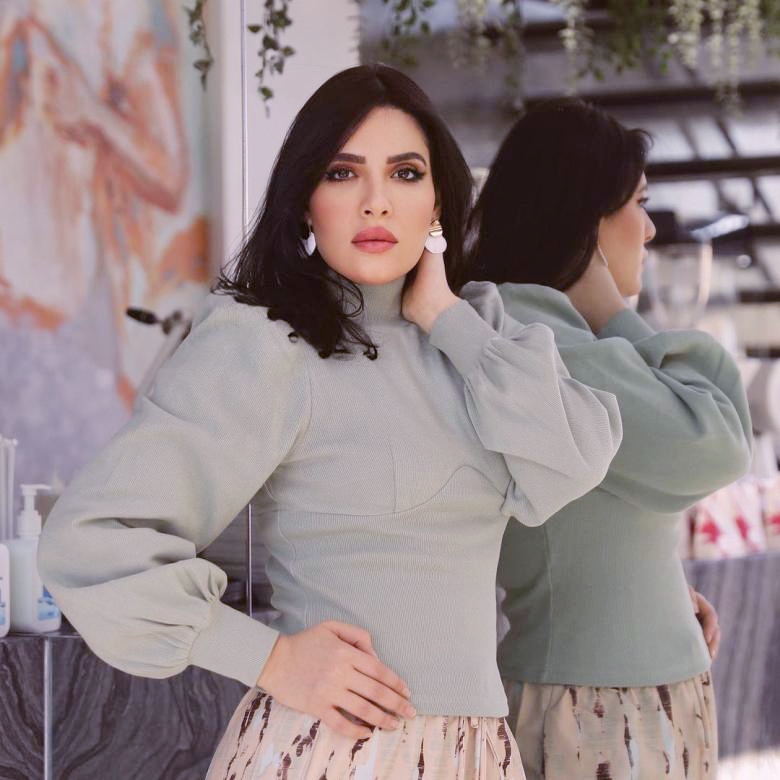 أزياء هبة الدري وهي واقفة أمام المرآة مرتدية كنزة خضراء وتنورة بنية 