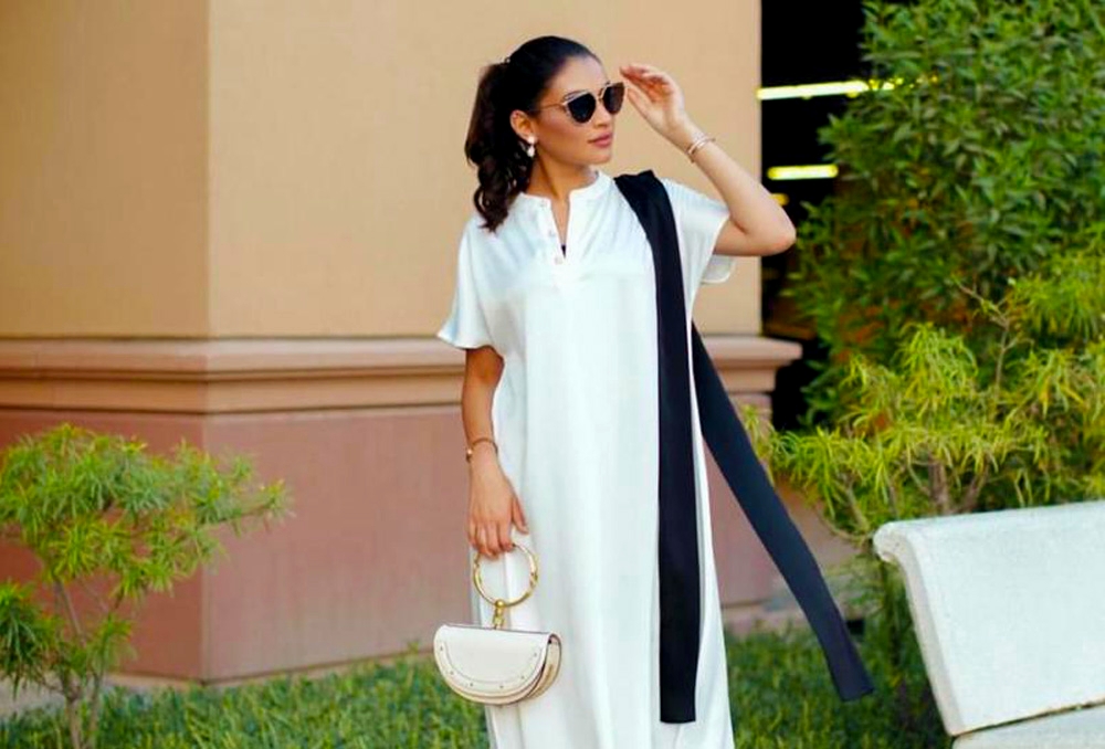 الفنانة شوق الهادي بفستان أبيض مع نظارات سوداء وتحمل حقيبة نصف دائرية لها سوار ذهبي